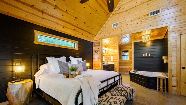 Een houten slaapkamer met een bed, nachtkastje, bankje en een badkamer met bad.