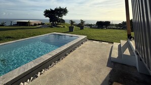 Una piscina con vista despejada al océano, rodeada de césped y algunos árboles.