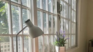 Un bureau en bois équipé d’un port USB, d’une lampe et d’une plante en pot aux fleurs violettes, placé près d’une fenêtre à barreaux.