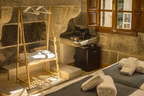 A stone-walled room with a bed, a small sink, and a window with wooden shutters. - NÓMADE SANTIAGO (Santiago de Compostela)