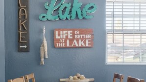 A dining area with a wooden table and chairs, a wall-mounted sign reading "Lake," and a smaller sign stating "Life is better at the lake."