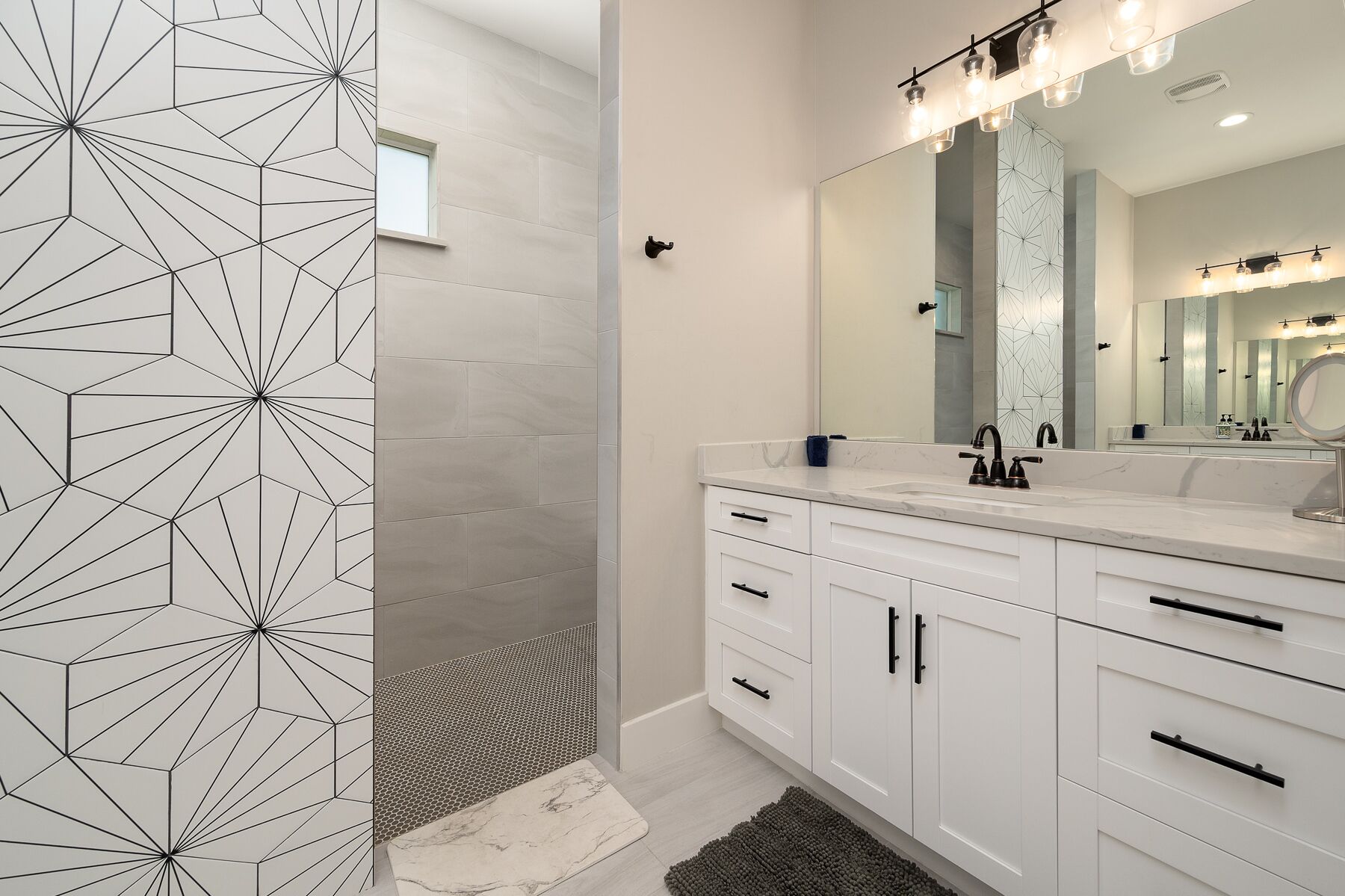 Une salle de bain moderne avec un mur de douche orné d’un motif géométrique, un grand miroir et un meuble-lavabo blanc doté de nombreux tiroirs.