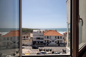 A coastal view from a window overlooking a street with buildings and parked cars. - Muito Acolhedor com Vista de mar (Colares)