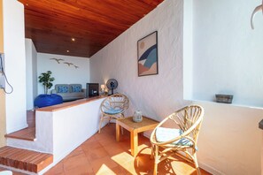 Ein sonnendurchfluteter Raum mit Holzbalkendecke, einer weißen Wand mit strukturierter Oberfläche, einem gerahmten abstrakten Gemälde, einem Korbstuhl mit Kissen, einem kleinen Tisch und einem blauen Sitzsack.