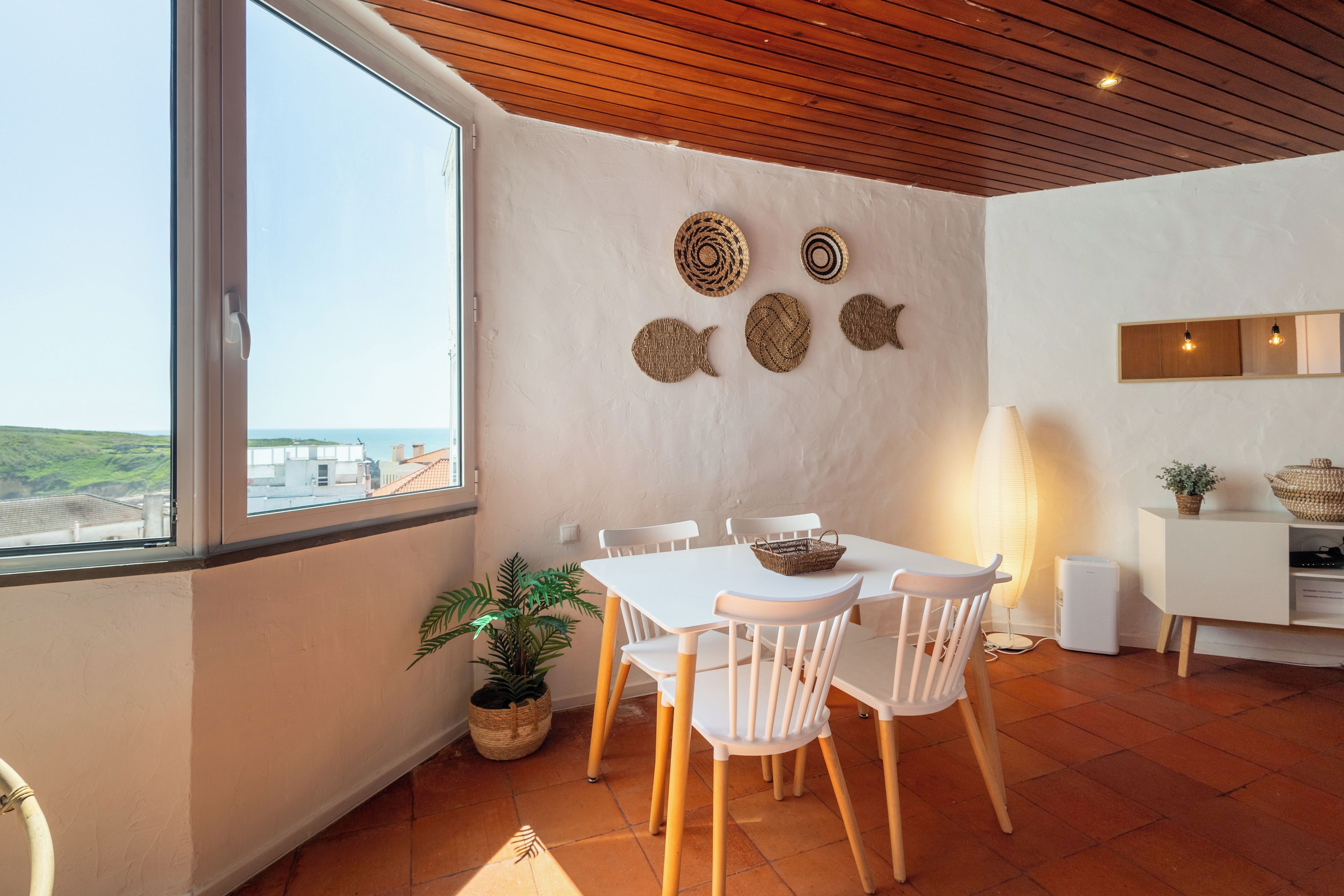 Een eethoek met een tafel en stoelen, een uitzicht door een groot raam, en een wand met decoratieve geweven objecten.