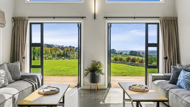 A spacious living room with two sofas, a coffee table, and a potted plant. Large sliding glass doors lead to a lush green landscape.