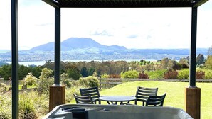 A hot tub with a table and chairs, surrounded by a wooden deck, overlooking a scenic landscape with mountains and a lake.