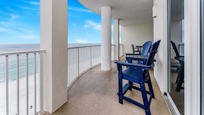 Ein Balkon mit zwei blauen Stühlen und einem Tisch, der Blick auf einen Strand mit blauem, wolkenlosem Himmel bietet.