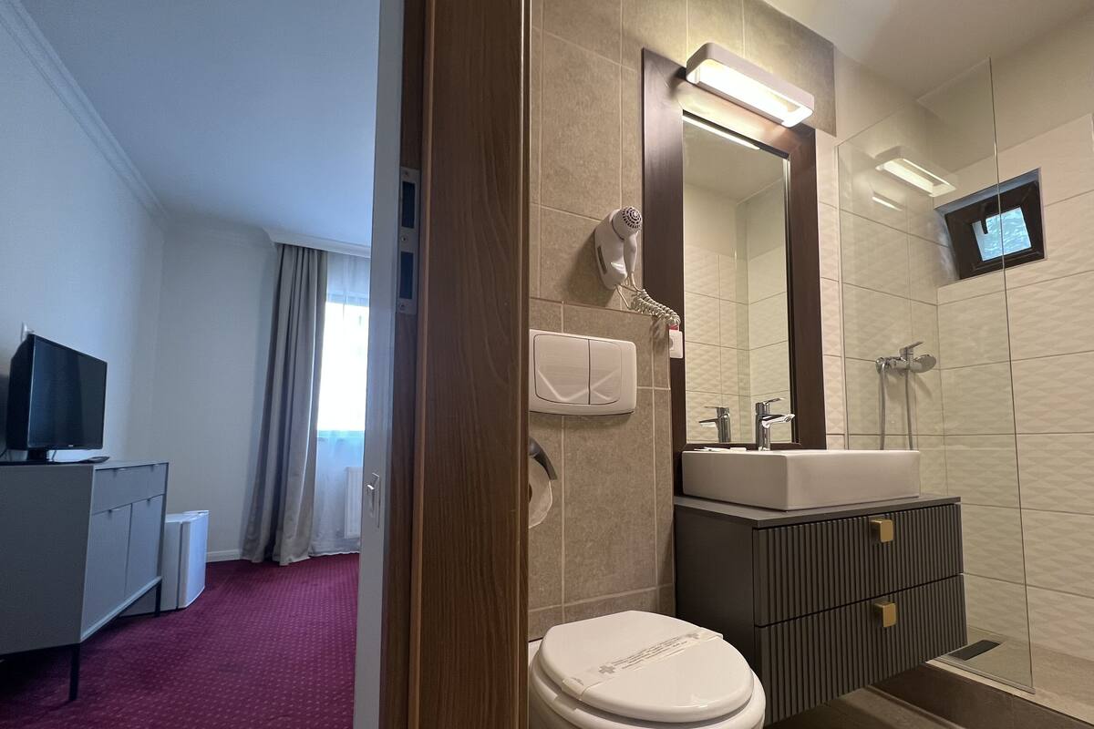 Un baño moderno con inodoro, lavamanos y ducha. Hay un televisor montado en la pared y un secador de pelo también en la pared.