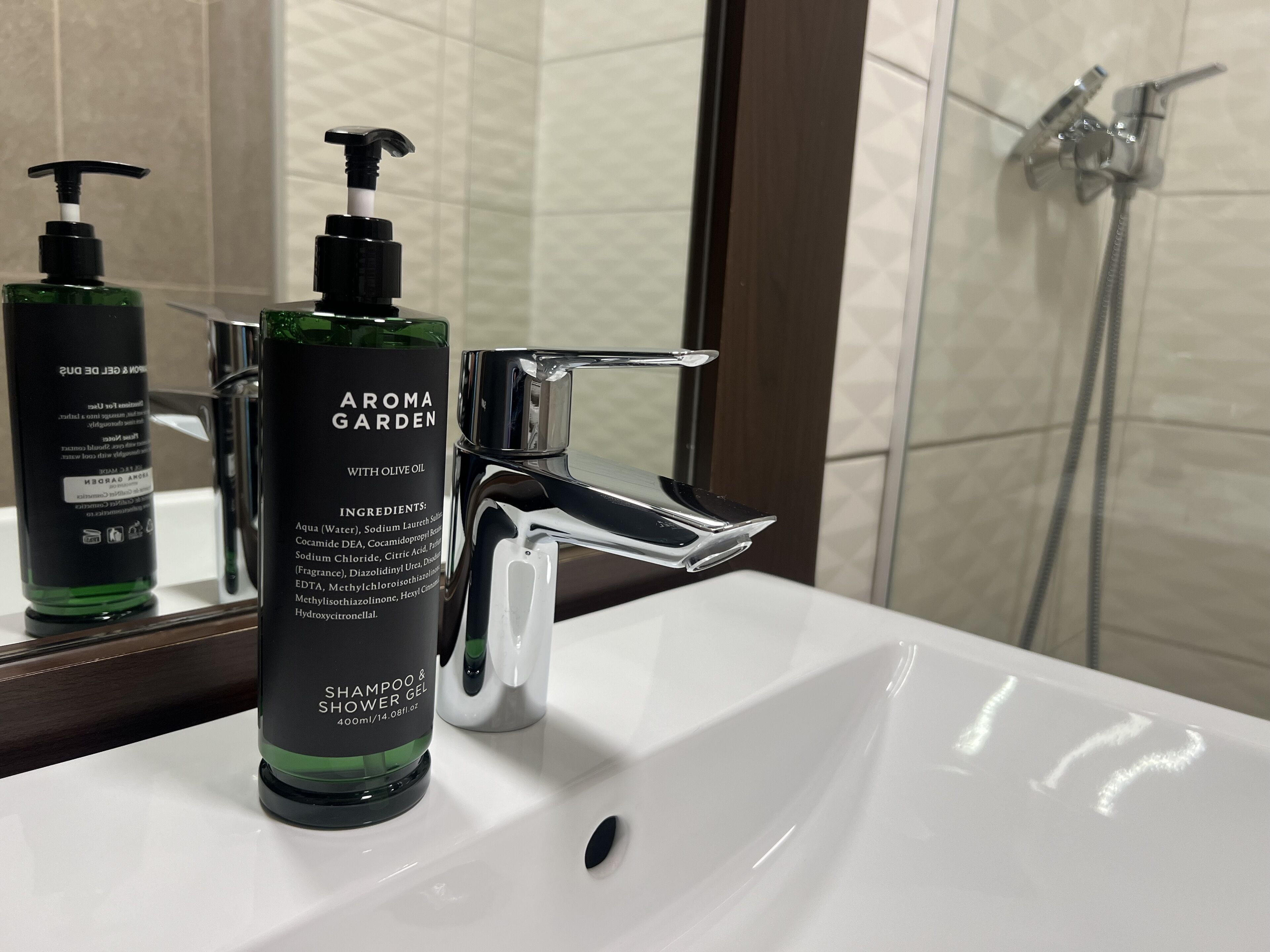Casa de banho com uma pia branca, um frasco verde de shampoo e gel de banho AROMA GARDEN, e uma torneira cromada.