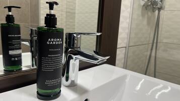 Casa de banho com uma pia branca, um frasco verde de shampoo e gel de banho AROMA GARDEN, e uma torneira cromada.