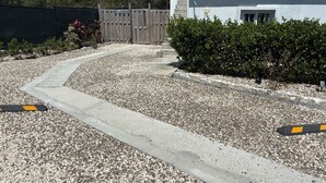 A gravel driveway leading to a white building with a wooden fence and a bush.