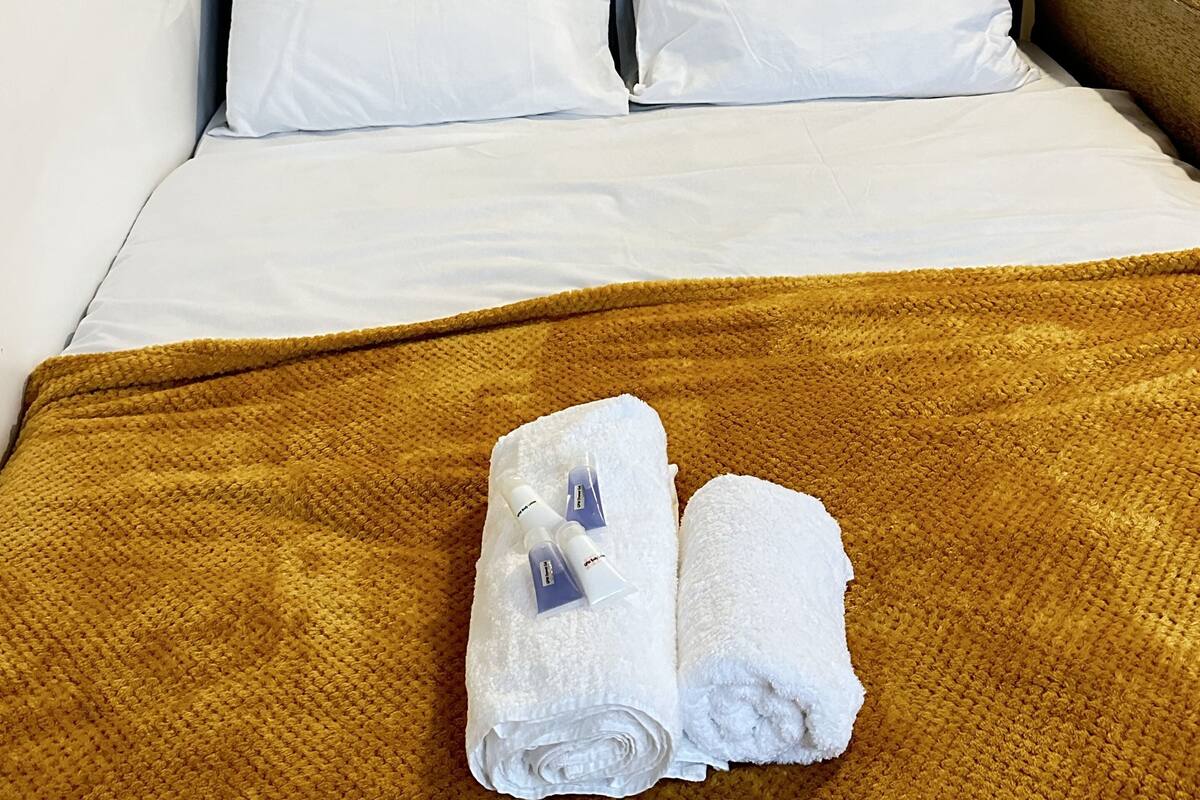 A neatly made bed with white linens, a brown textured blanket, and two rolled towels on the bed.
