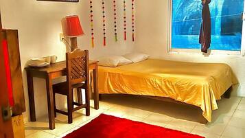 A bedroom with a bed, a desk with a chair, a red rug, a hanging decoration, and a window with a curtain.