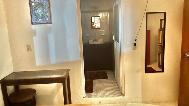 A small room with a bed, a table, a mirror, and a door leading to a bathroom. - Room-central Comfy Small Studio (San Cristóbal de las Casas)