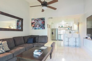 A modern living room with a sectional sofa, a coffee table, and a kitchen area with white cabinets and a stainless steel refrigerator.
