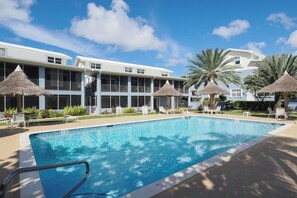 A swimming pool with lounge chairs and thatched umbrellas, surrounded by a building with balconies and a clear sky.