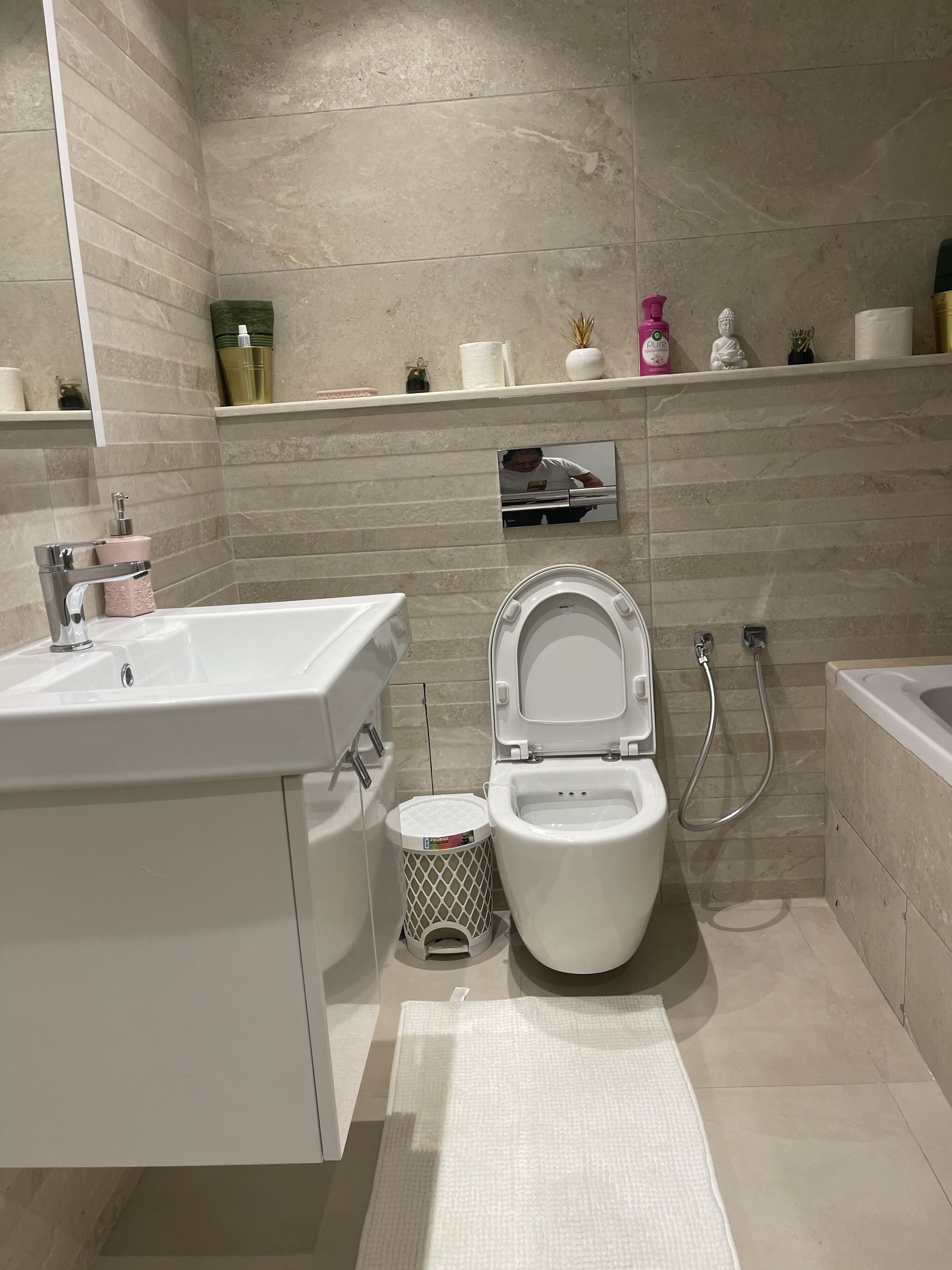 A modern bathroom with a white sink, toilet, and bathtub. There are shelves with toiletries and a small mirror.