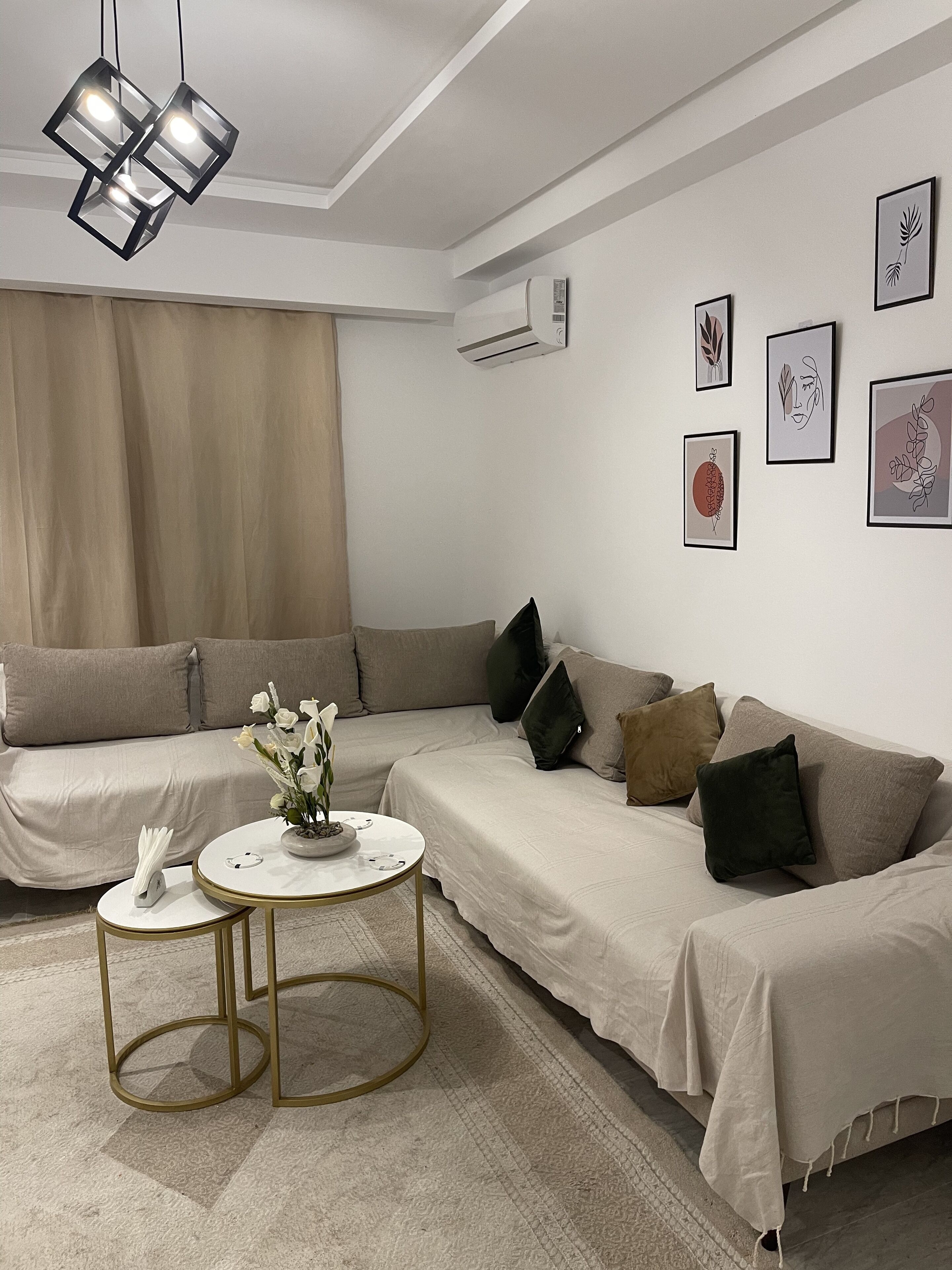 Ein modernes Wohnzimmer mit einer beigefarbenen Sofagarnitur, einem runden Couchtisch mit Blumenvase und schlichter Einrichtung mit gerahmten Kunstwerken an der Wand.