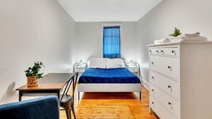 Un dormitorio con cama, mesitas de noche, cómoda, ventana con cortinas, una planta sobre una mesa y una silla.