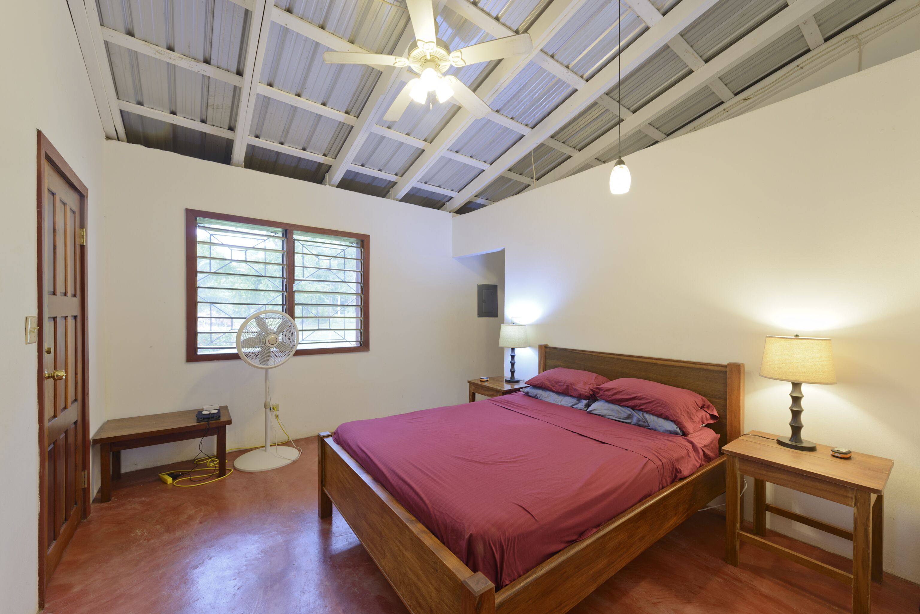 A bedroom with a wooden bed, a standing fan, a bedside lamp, a window with bars, and a ceiling fan.