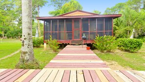 A wooden deck leading to a building with large windows and a red roof, surrounded by greenery. - Beautiful cabin nestled in tropical surroundings on the Banks of the Sitee River (Hopkins)