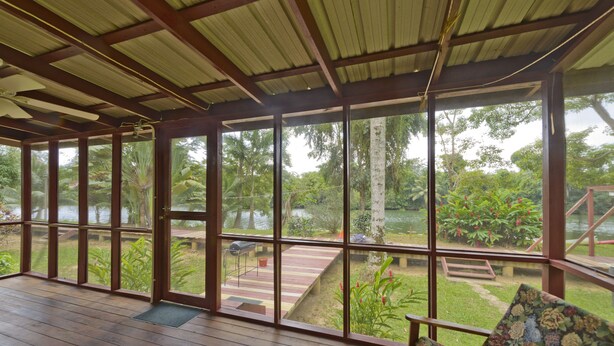 A covered outdoor area with wooden flooring, large windows, and a view of a garden and water body. - Beautiful cabin nestled in tropical surroundings on the Banks of the Sitee River (Hopkins)
