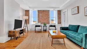 A living room with a brick wall, white walls, a green sofa, a wooden coffee table, two white chairs, a TV on a wooden stand, and a small table with a plant. - Uncle Sam's Hideaway |Nearby Colleges & Hospital|Personal Workspace|Pet Friendly (Troy)