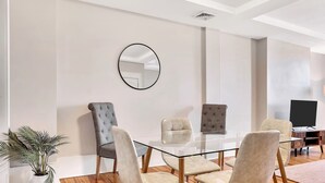 A modern dining area with a glass table, four upholstered chairs, a potted plant, and a television in the background. - Uncle Sam's Hideaway |Nearby Colleges & Hospital|Personal Workspace|Pet Friendly (Troy)