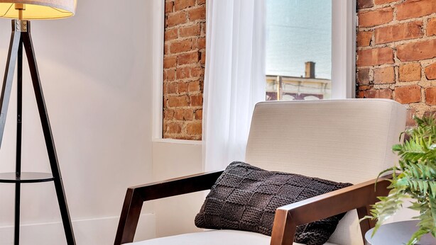 A chair with a knitted pillow, a floor lamp, and a potted plant in a room with a brick wall and a window. - Uncle Sam's Hideaway |Nearby Colleges & Hospital|Personal Workspace|Pet Friendly (Troy)