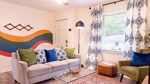 A living room with a sofa, armchair, glass coffee table, patterned rug, and a wall with a colorful abstract design. - Artist's Bungalow- 1/2 mi from Covington Square (Covington)