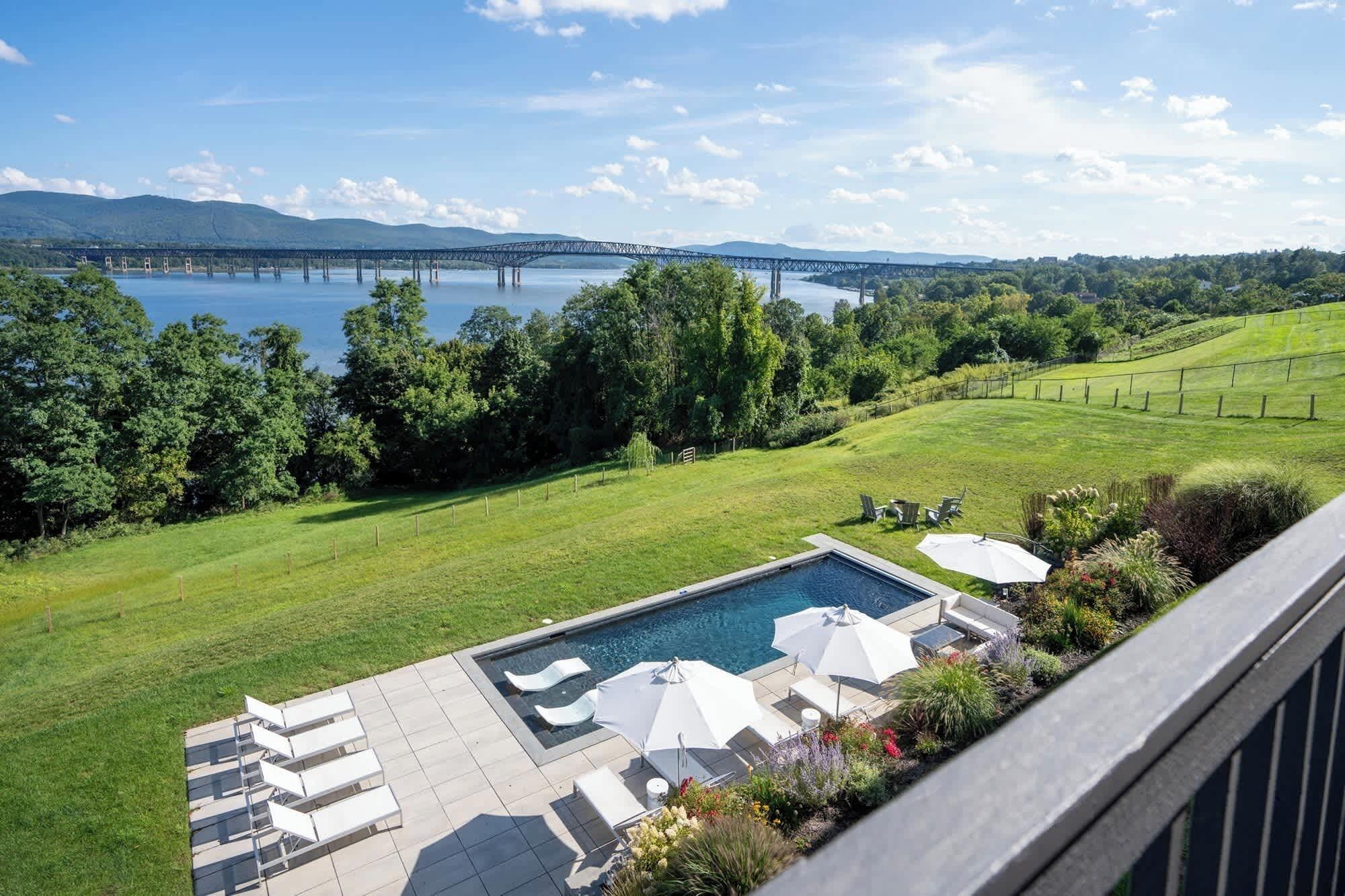 A pool area with lounge chairs and umbrellas, a view of a bridge over a lake, and a grassy landscape with trees.