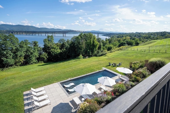 A pool area with lounge chairs and umbrellas, a view of a bridge over a lake, and a grassy landscape with trees.