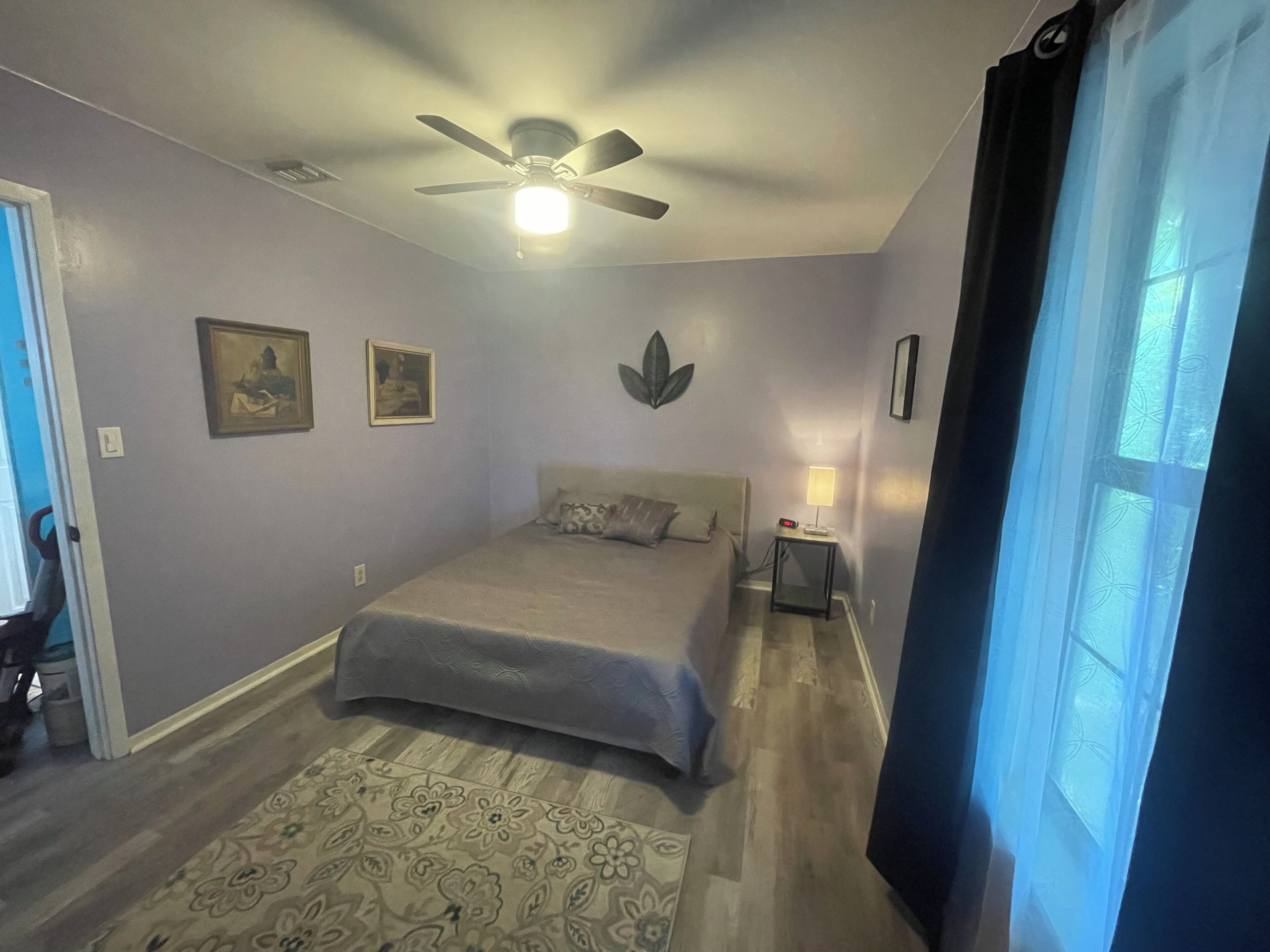 A bedroom with a bed, a ceiling fan, a nightstand with a lamp, and a window with curtains.