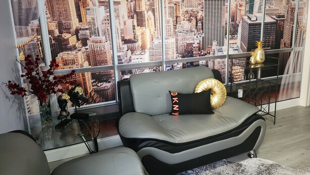 A modern living room with a sofa and a glass coffee table, a decorative vase with red flowers, and a large window with a cityscape view.