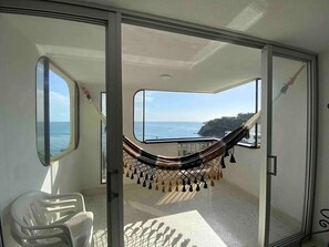 Een kamer met een hangmat bij een groot raam met uitzicht op de oceaan, een stoel en een zicht op een rotsachtige kustlijn.