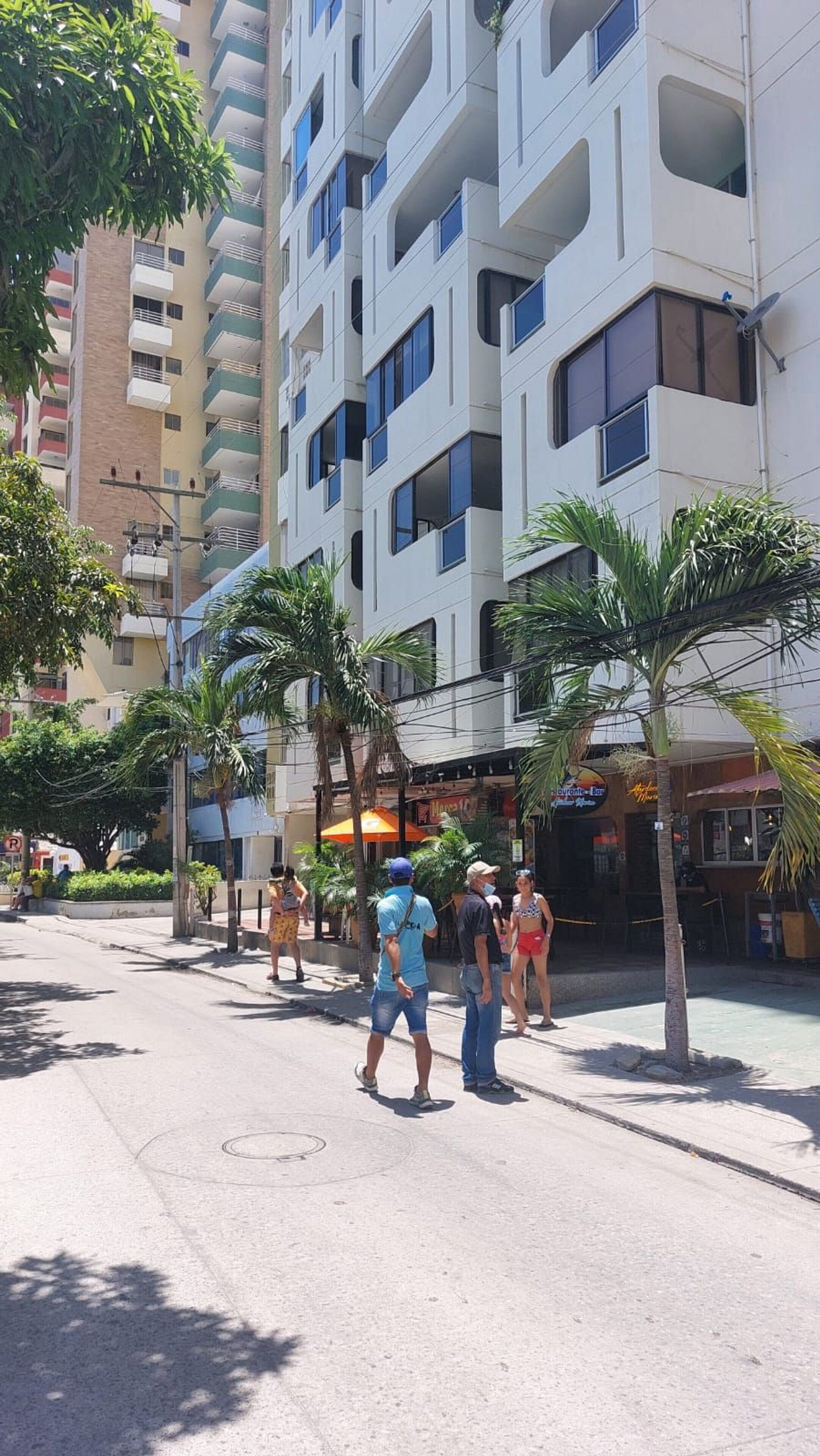 A multi-story building with balconies, a street with pedestrians, and palm trees.