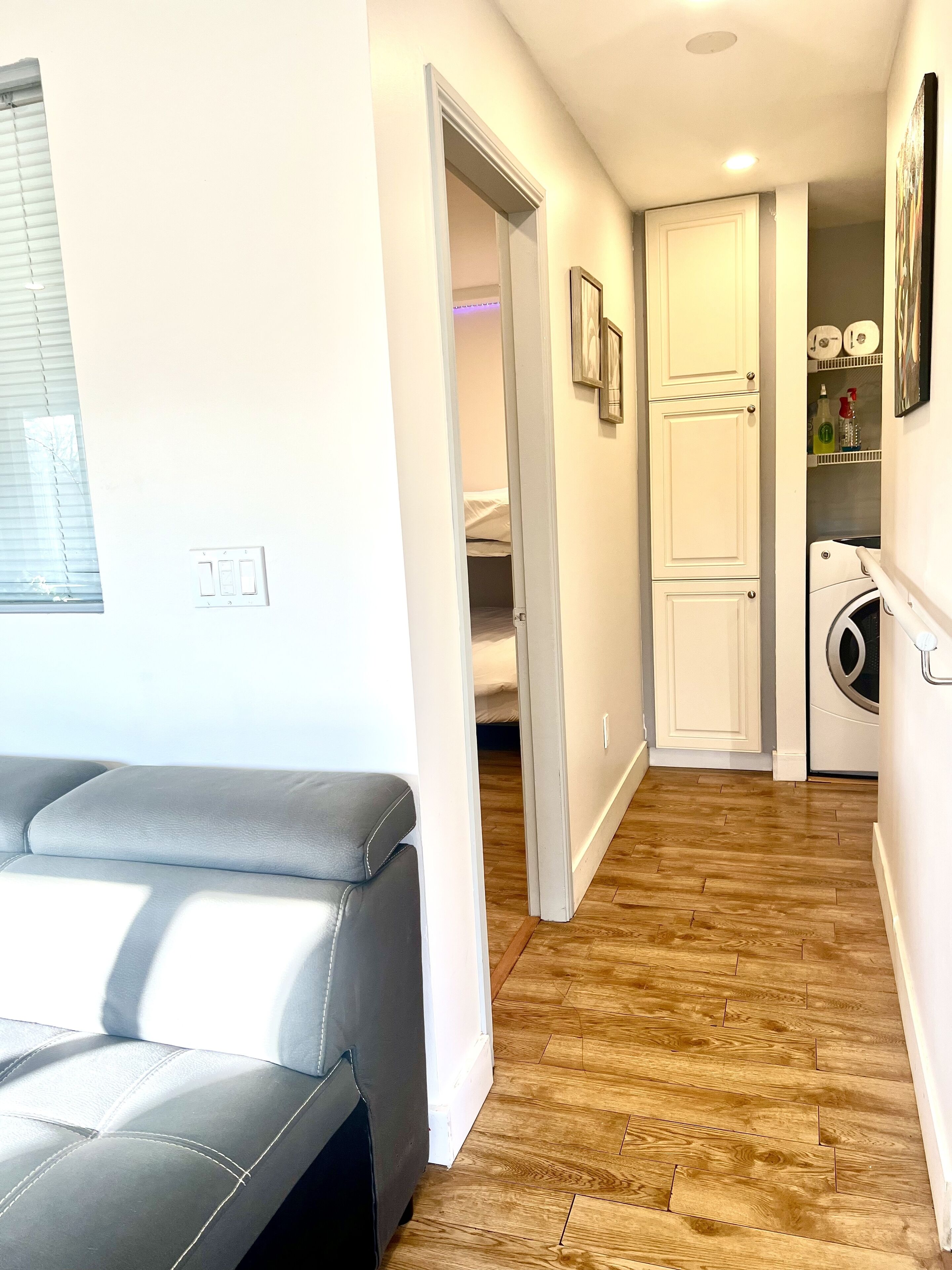 A modern apartment with a grey sofa, wooden flooring, a laundry area, and a door leading to a bedroom.