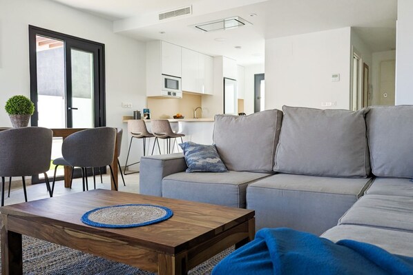 A modern living room with a grey sofa, a wooden coffee table, and a dining area with chairs and a small kitchen in the background.