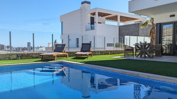 A modern pool area with two lounge chairs, a dining table with chairs, and a view of the city skyline. - PANORAMIC pool & mountains house (Finestrat)