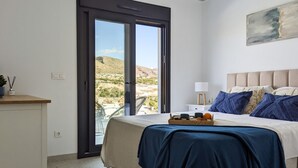 A bedroom with a bed, a nightstand, a lamp, a window with a view of the countryside, and a painting on the wall. - PANORAMIC pool & mountains house (Finestrat)