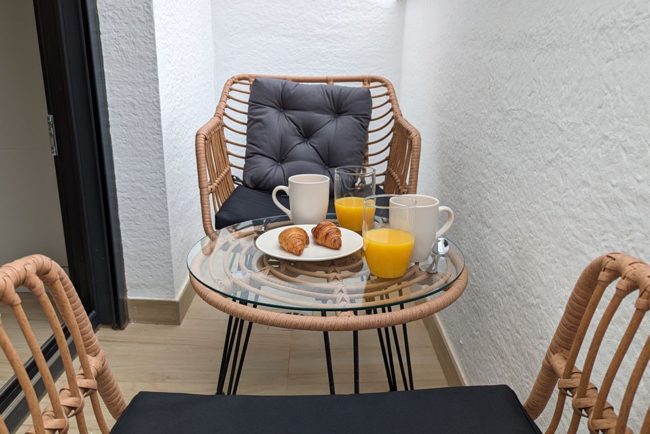 Une table en verre, placée dans un coin, est dressée pour le petit-déjeuner avec des croissants, du jus d’orange et du café, et entourée de chaises en osier.