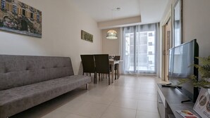 Ein Wohnzimmer mit grauem Sofa, einem Esstisch mit Stühlen, einem Fernseher und einem Fenster mit Vorhängen.