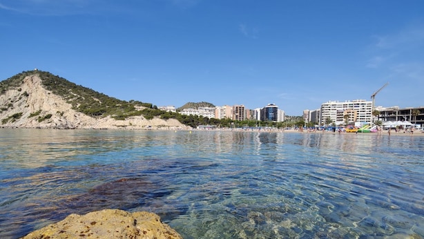 Eine Küstenlandschaft mit Strand, klarem Wasser, felsigem Ufer und Gebäuden im Hintergrund.