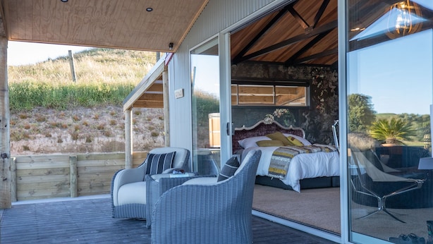 A bedroom with a large bed, two wicker chairs, and a glass door leading to a balcony. - Holiday Bliss - Tirau, Piarere (Piarere)