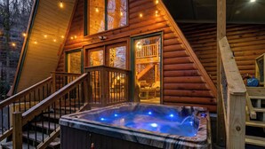 A wooden cabin with a hot tub, wooden stairs, and string lights.