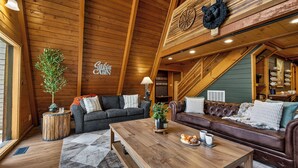 A cozy cabin interior with a wooden ceiling, a fireplace, a sofa, a coffee table, and a dining area.