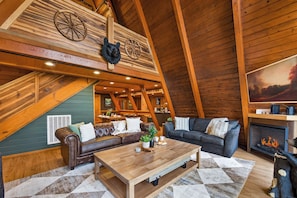 Een gezellige loft woonkamer met een open haard, leren banken, een houten salontafel en een hoge plafond met zichtbare balken.