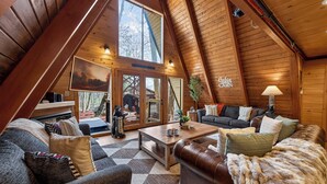 A cozy cabin interior with a high ceiling, wooden walls, a large window, and a fireplace.