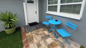 Un pequeño patio con una puerta blanca, una ventana con persianas y una mesa y sillas azules.
